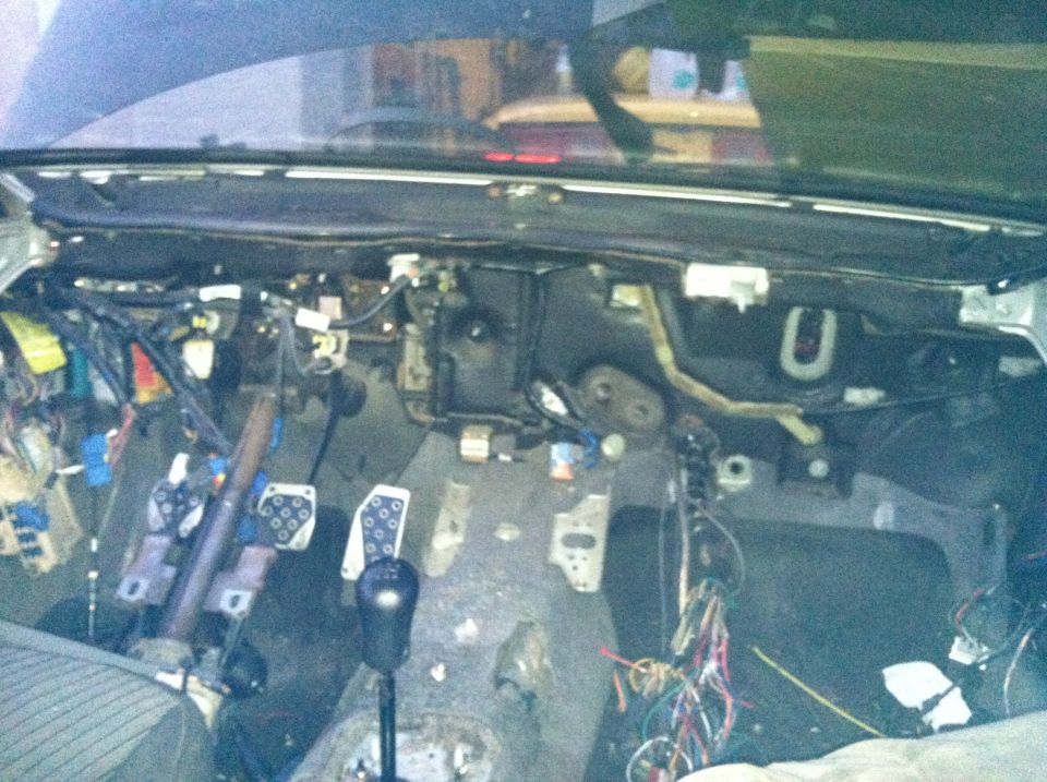 Car interior showing dashboard removed, exposed wires, and pedals. Looks like a manual transmission