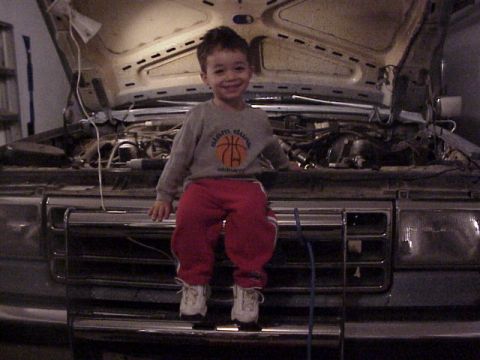1989 Ford Bronco - A young boy smiles while sitting on the front grille of a 1989 Ford Bronco with its hood open, revealing the engine.