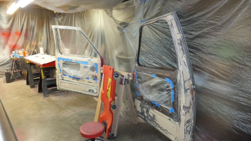 Two vehicle doors, covered in plastic and tape, are propped up in a workshop. One door is on sawhorses, the other is on a stand