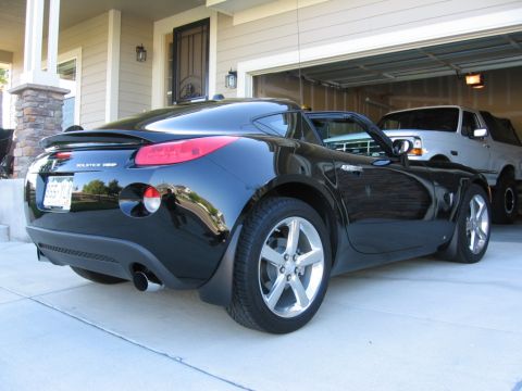 1995 Ford Bronco - A black Pontiac Solstice GXP Coupe is parked outside a garage, with a white pickup truck visible inside.