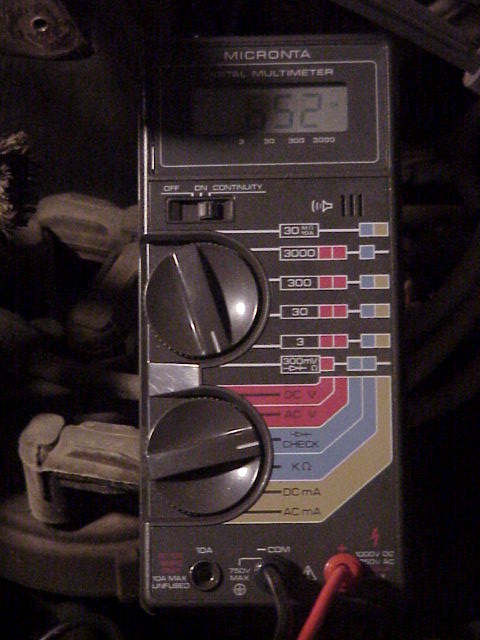 1989 Ford Bronco - A Micronta digital multimeter held over a dark engine bay, its LCD reading roughly 12.52 with the rotary selector set for DC volts and red…