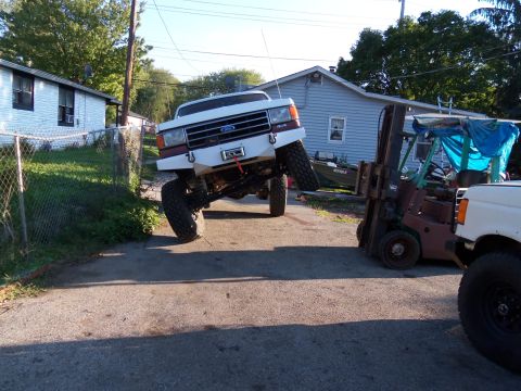 1989 Ford Bronco - White Ford truck with lifted suspension and aftermarket bumper is angled upwards, with a forklift nearby.