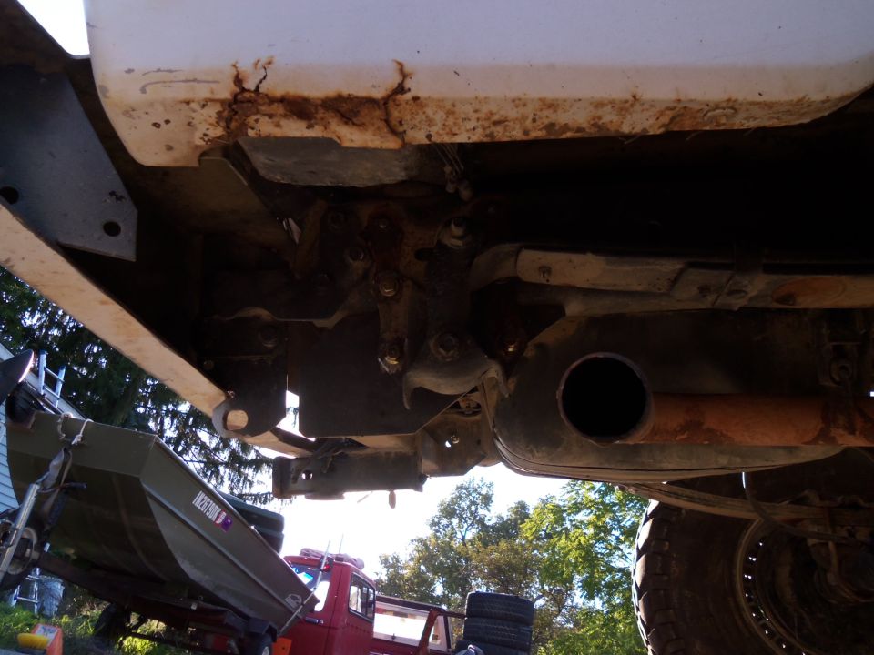 Underside view of vehicle rear suspension components, exhaust pipe, and muffler. Some rust is visible on the frame