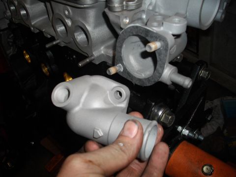 1989 Ford Probe - A hand holds a gray thermostat housing, preparing to install it onto an engine block.