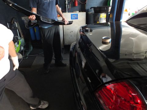 2003 Ford Crown Victoria - person carries a black spoiler trim toward the rear deck of a black sedan inside a repair shop.