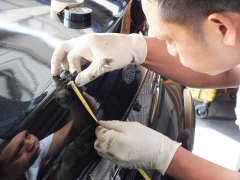 2003 Ford Crown Victoria - worker leans over a black sedan's trunk, marking a measurement with a yellow tape measure.