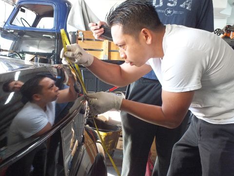 2003 Ford Crown Victoria - worker measures along a black car's body with a tape measure, an old truck visible in the garage.