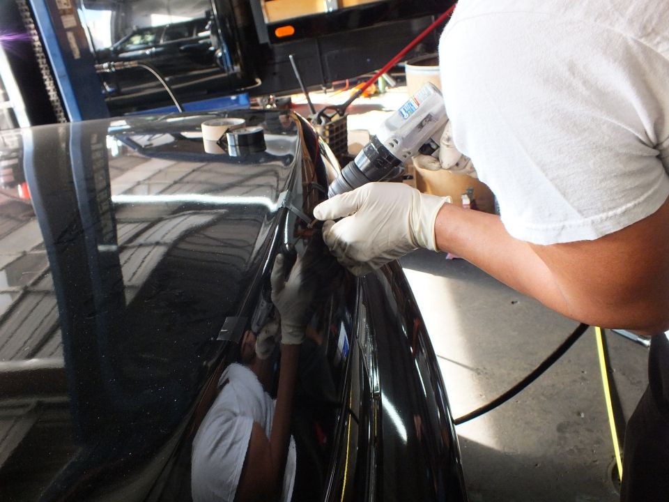 Gloved worker using power drill along trunk edge of black car under shop lighting