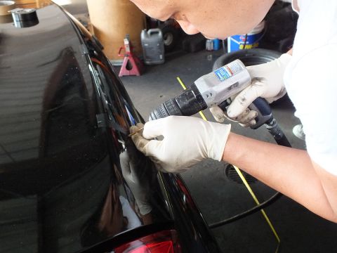2003 Ford Crown Victoria - worker drills into the trunk edge of a black sedan, masking tape and tools surrounding the work area.