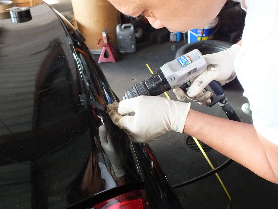 Gloved hands drilling trunk edge of black sedan with red taillight in garage