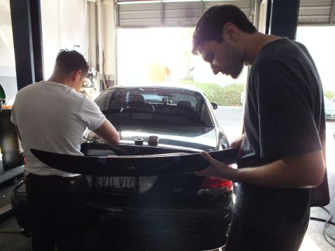 2003 Ford Crown Victoria - Garage scene as two technicians fit a trunk-mounted wing to a dark four-door car.