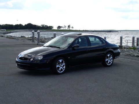 1998 Ford Taurus - This black 1998 Ford Taurus is captured from a three-quarter front view, parked beside a body of water with a scenic background.