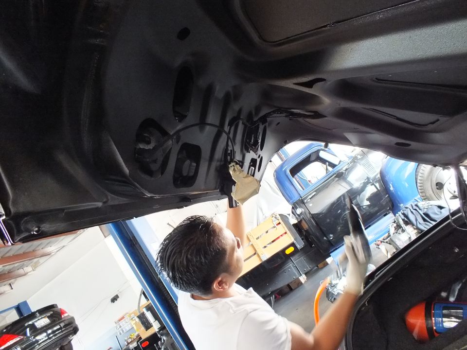 Man in gloves works under open black trunk lid inside a vehicle shop bay