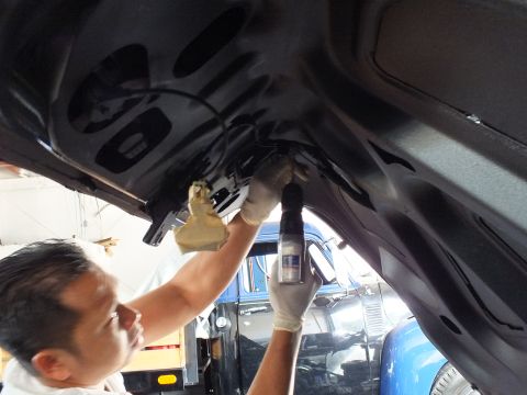 2003 Ford Crown Victoria - Worker in latex gloves sprays the inner trunk panel of a dark vehicle raised on a lift, blue truck behind