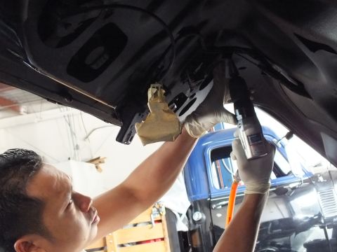 2003 Ford Crown Victoria - Gloved hands work a spray tool against the underside of a dark car panel, vintage blue truck cab behind