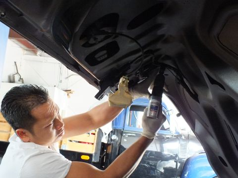 2003 Ford Crown Victoria - Man in white shirt and gloves sprays inner panel of raised dark vehicle, blue truck and shop racks behind