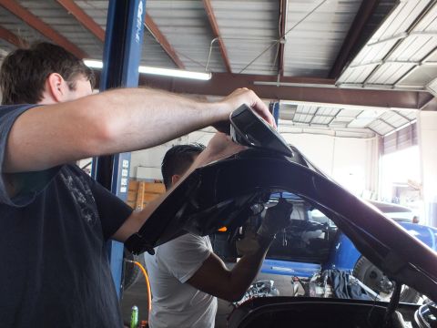 2003 Ford Crown Victoria - One man holds a panel while another works beneath the open hood of a dark car, blue vehicle behind