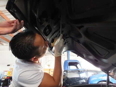 2003 Ford Crown Victoria - Gloved technician handles a tool beneath the open decklid of a dark vehicle on a lift, blue truck behind