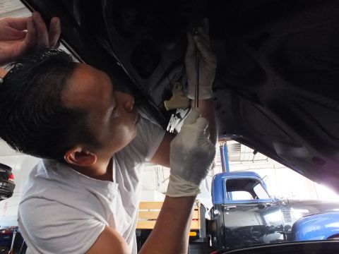 2003 Ford Crown Victoria - Worker in gloves grips pliers against the underside of a dark car panel, blue truck visible in background