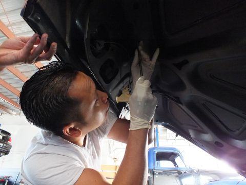 2003 Ford Crown Victoria - Technician positions a rod into the underside of a dark car's open panel, blue truck cab behind
