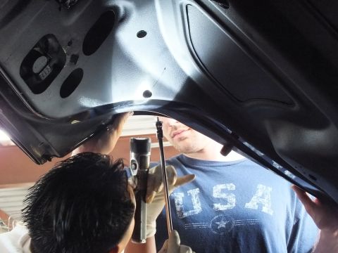 2003 Ford Crown Victoria - Two technicians look at the underside of a dark car's open trunk lid, lifted vehicles behind in the shop