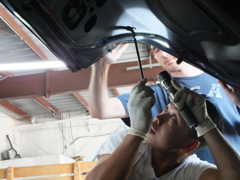 2003 Ford Crown Victoria - Gloved worker guides a support rod into a dark car panel as a teammate in a USA shirt shines a light