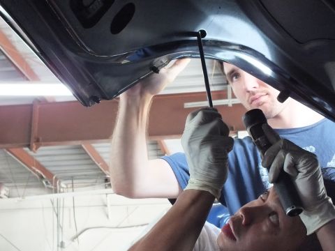 2003 Ford Crown Victoria - One technician inserts a strut into the inner panel as a helper holds a flashlight beneath the decklid
