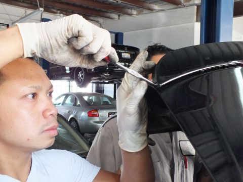 2003 Ford Crown Victoria - Worker uses a flat tool on a black car's trunk; a silver sedan sits in the shop behind