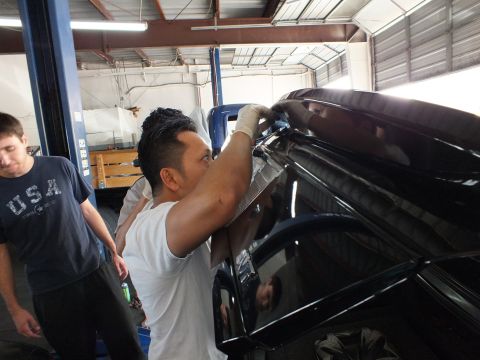 2003 Ford Crown Victoria - Worker pulls trim off a black sedan; a second man and old blue pickup stand behind