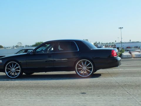 2003 Ford Crown Victoria - Profile of a lowered black sedan rolling on big chrome wheels past warehouses
