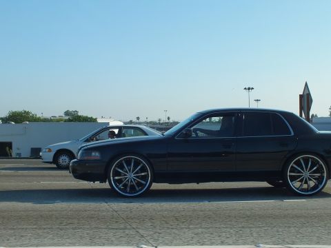 2003 Ford Crown Victoria - Side profile of a black sedan on large chrome wheels passing a white sedan on the road