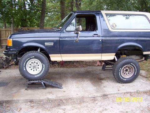1990 Ford Bronco - A dark blue 1990 Ford Bronco is shown from the side, lifted on jack stands with the front wheel removed for maintenance.