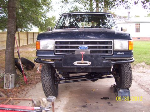 1990 Ford Bronco - A dark blue Ford truck is shown from the front, lifted on blocks with a winch bumper installed.