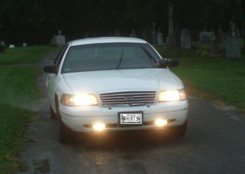 1998 Ford Crown Victoria - A white 1998 Ford Crown Victoria is parked in a cemetery at dusk, its headlights illuminating the road.