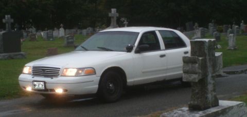 1998 Ford Crown Victoria - A white 1998 Ford Crown Victoria is parked in a cemetery, with tombstones visible in the background.