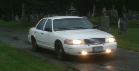 1998 Ford Crown Victoria - A white sedan drives down a path in a cemetery, surrounded by gravestones.