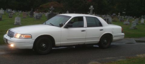 1998 Ford Crown Victoria - A white 1998 Ford Crown Victoria sedan is parked on a wet road in a cemetery.