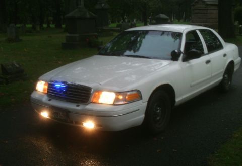 1998 Ford Crown Victoria - White 1998 Ford Crown Victoria police car with blue and white lights on, parked in a cemetery.