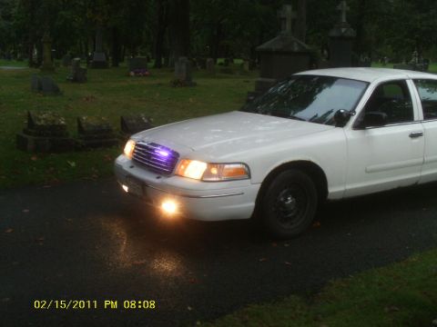 1998 Ford Crown Victoria - A white sedan sits in a cemetery at dusk, its lights illuminating the wet pavement.