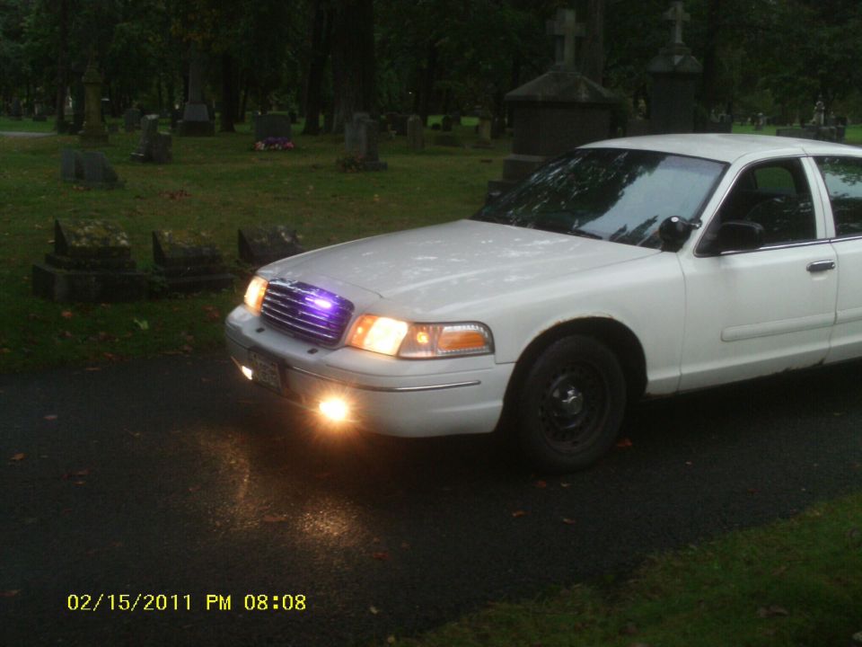 White sedan parked on asphalt in a cemetery with tombstones in background. Headlights and grille lights are on