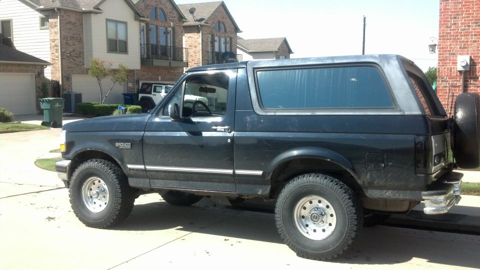 Black Ford Bronco SUV, side view, lifted on alloy wheels with rear spare tire, parked in a suburban driveway