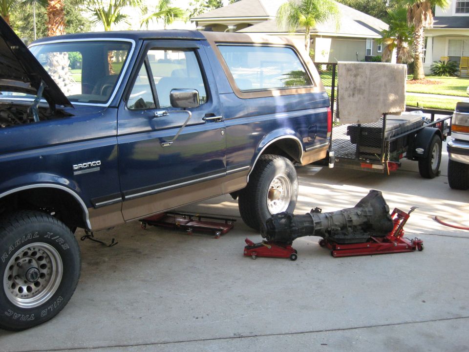 Blue Ford Bronco with hood open, transmission removed, on jacks. Trailer behind