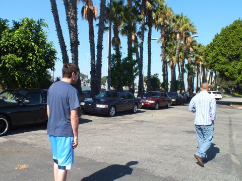 2003 Ford Crown Victoria - Man in gray shirt and blue shorts and another in striped shirt cross a sunlit lot of parked cars
