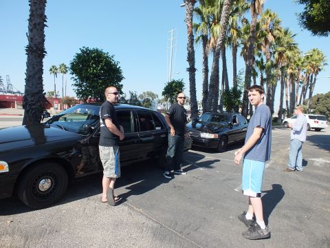 2003 Ford Crown Victoria - Several men gather around a black full-size sedan with industrial cranes in the background