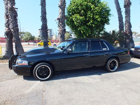 2003 Ford Crown Victoria - Lowered black sedan parked broadside before a green tree and industrial silos