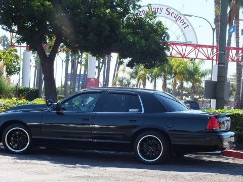 2003 Ford Crown Victoria - Lowered black sedan on black wheels angled before the red Queen Mary Seaport arch