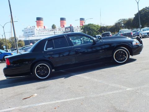 2003 Ford Crown Victoria - Dark sedan on black wheels parked broadside, red and black ship funnels in the distance