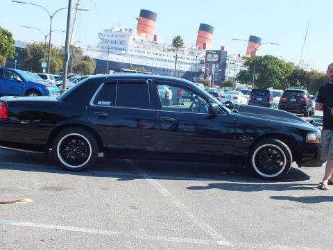 2003 Ford Crown Victoria - Dark sedan on black wheels parked broadside as a man looks on, ship funnels behind