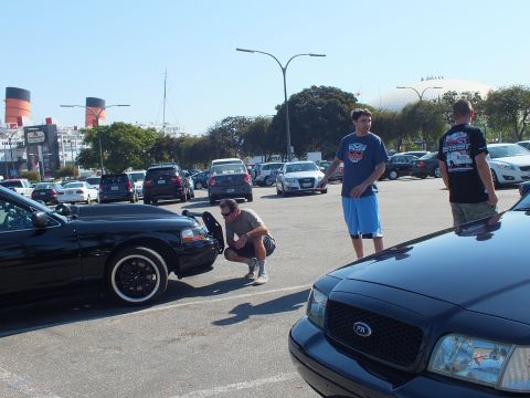 2003 Ford Crown Victoria - Person crouches at a sedan's front fender while others stand between cars near the ship