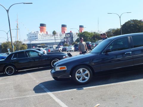 2003 Ford Crown Victoria - Two dark sedans face opposite ways in a parking lot, red-and-black ship funnels rising past streetlights behind them.